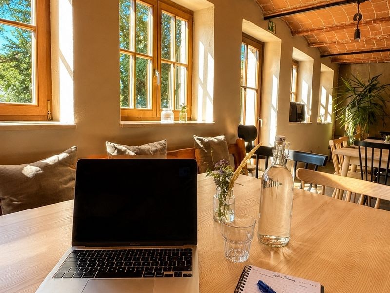 Laptop und Notizbuch auf dem Holztisch im Stall, Sonnenlicht durch die alten Fenster
