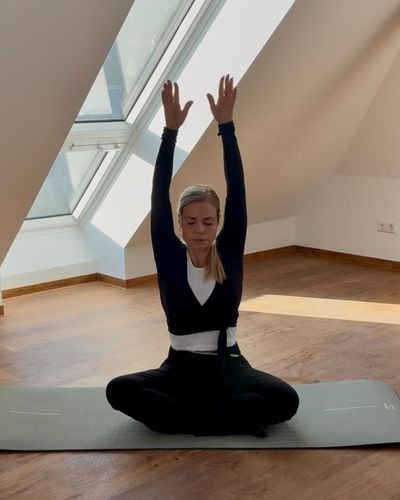 Yogalehrerin Tamara sitzt mit erhobenen Armen und geschlossenen Augen auf ihrer Yogamatte